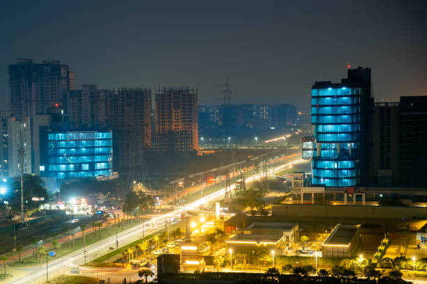 Aerial night not of gurgaon with light trails on the road and blue lights on the building. Shows the cityscape with bustling roads, towers with offices and residences and construction sites for new real estate projects